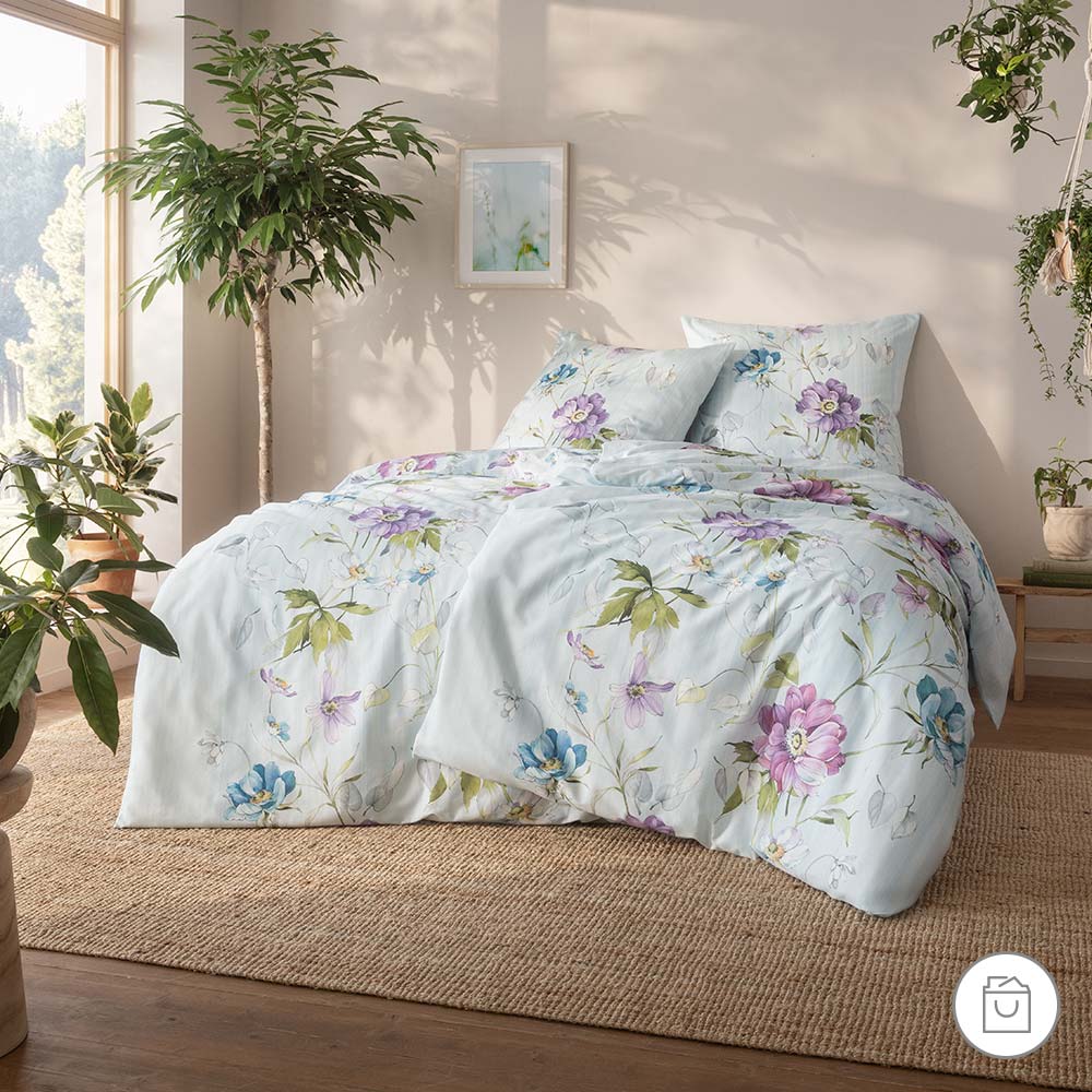 Bio-Bettwäsche mit floralen Aquarellblumen in Violett, Blau und Grün auf hellem Hintergrund in heller Schlafzimmerumgebung.