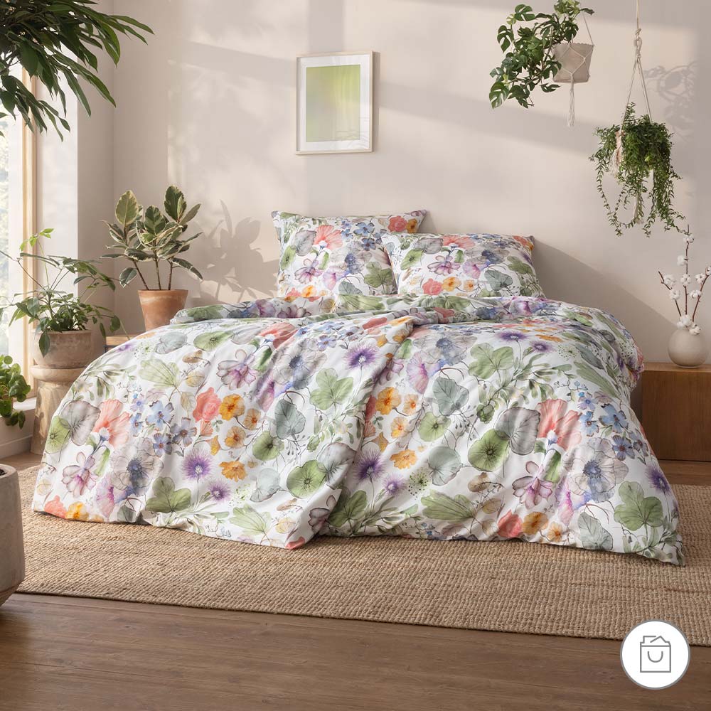 Bio-Bettwäsche mit buntem Blüten- und Blättermuster auf hellem Grund in gemütlicher Schlafzimmerumgebung.