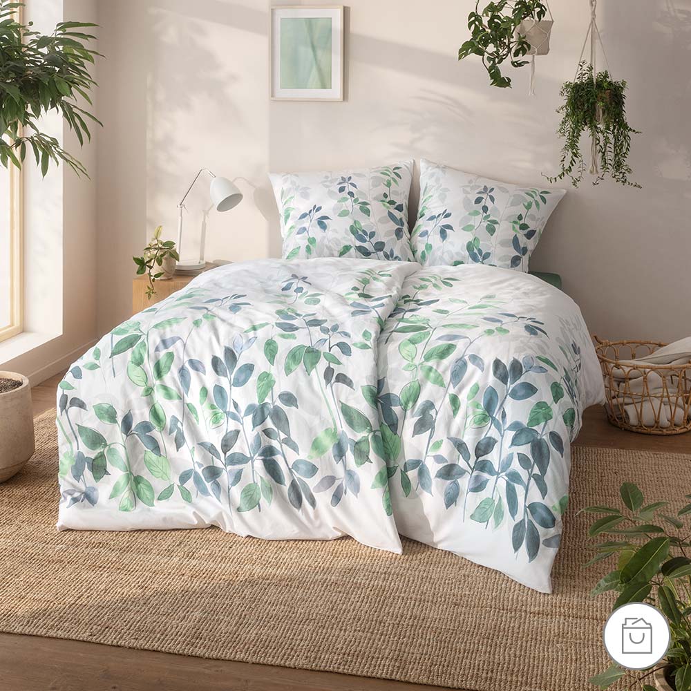 Bio-Bettwäsche mit grün-blauem Blättermuster auf weißem Hintergrund in heller Schlafzimmerumgebung.