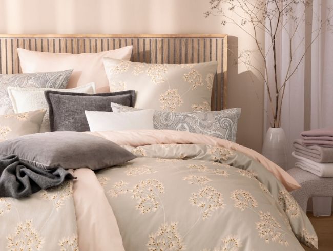 Bettwäsche Antje mit zartem Blütenmotiv in Beige, kombiniert mit unifarbener Bettwäsche Valetta, verschiedenen Dekokissen und Wohndecken auf Doppelbett in hellem Schlafzimmer