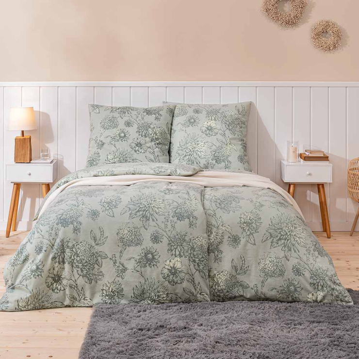 Bettwäsche Luserna mit großen Chrysanthemenblüten und detailreichen Blätterzeichnungen in gedämpften Grün- und Grautönen auf graugrünen Grund auf Bett im Schlafzimmer