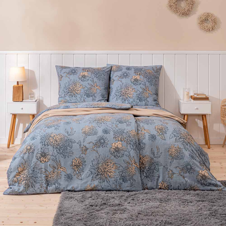 Bettwäsche Luserna mit großen Chrysanthemenblüten und detailreichen Blätterzeichnungen in Blau-, Beige- und Grautönen auf blaugrauem Grund auf Bett im Schlafzimmer