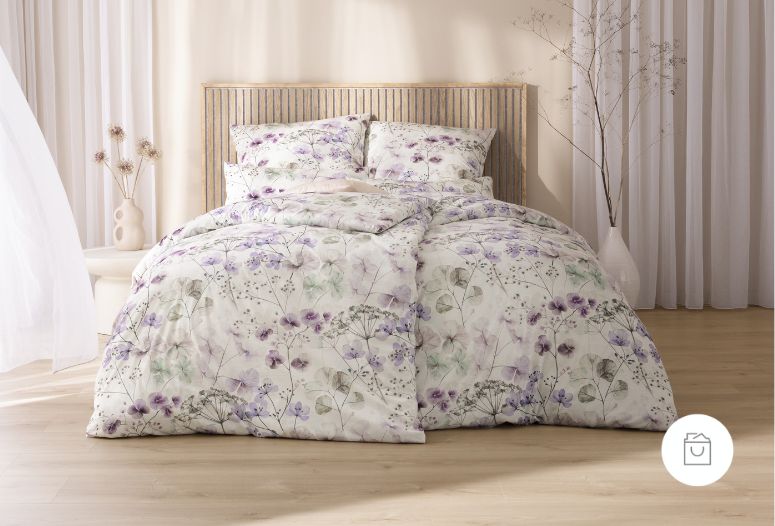 Bettwäsche Sonja mit feinen Wiesenblumen, zarten Blüten und filigranen Blättern in Violett-, Grün- und Cremetönen auf hellem Grund auf Bett im Schlafzimmer