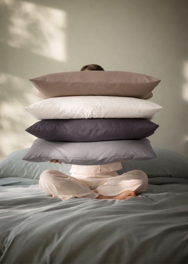 Person sitzt auf Bett mit Mako-Satin-Bettwäsche Valetta und hält einen Stapel Kissen in Kisel, Weiss, Anthrazit und Platin vor dem Gesicht.