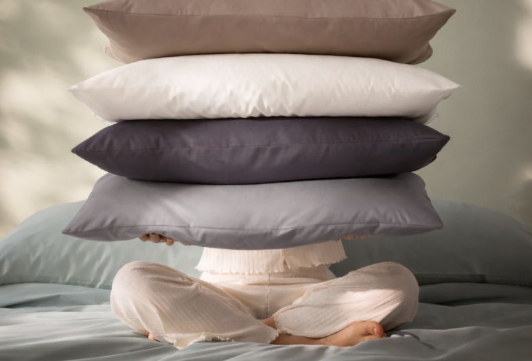 Person sitzt auf Bett mit Mako-Satin-Bettwäsche Valetta und hält einen Stapel Kissen in Kisel, Weiss, Anthrazit und Platin vor dem Gesicht.