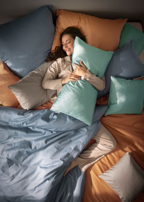 Frau liegt lächelnd im Bett mit Mako-Satin-Bettwäsche Valetta in verschiedenen Farben, umgeben von mehreren Kissen in Blau-, Grau- und Apricottönen.