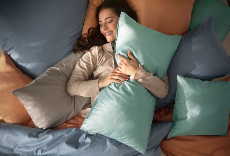 Frau liegt lächelnd im Bett mit Mako-Satin-Bettwäsche Valetta in verschiedenen Farben, umgeben von mehreren Kissen in Blau-, Grau- und Apricottönen.