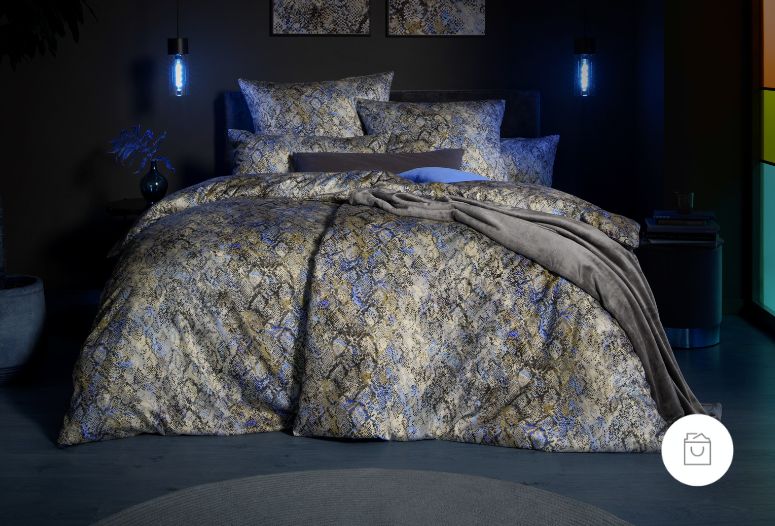 Bettwäsche aus Mako-Satin mit modernem Schlangenprint in Gold-, Beige- und Blautönen auf dunklem Hintergrund – inszeniert in stimmungsvoller Abendbeleuchtung.