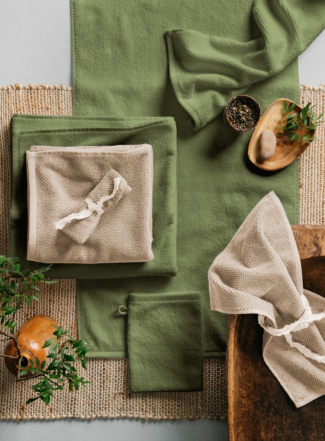 Bio-Frottierwaren in Beige und Grün auf Naturfasermatte mit dekorativen Accessoires arrangiert. 