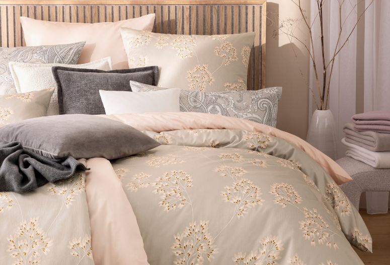 Bettwäsche Antje mit zartem Blütenmuster in Beige und Taupe, kombiniert mit unifarbener Bettwäsche Valetta, dunkler Wohndecke Trevi und verschiedenen Dekokissen Tokio und Velvet auf Doppelbett in hellem Schlafzimmer