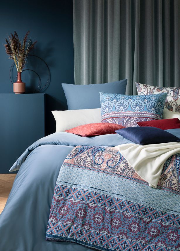Bettwäsche-Arrangement auf modernem Doppelbett: Paisley-Bettwäsche Carmen, unifarbene Bettwäsche Valetta, helles Paisley-Kissen Saskia im Hintergrund sowie verschiedene Dekokissenbezüge und Wohndecken in Blau-, Creme- und Rottönen.
