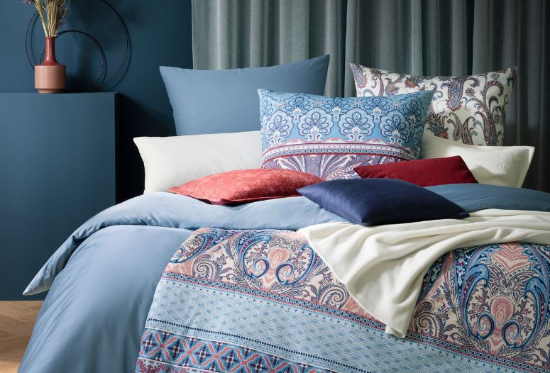 Bettwäsche-Arrangement auf modernem Doppelbett: Paisley-Bettwäsche Carmen, unifarbene Bettwäsche Valetta, helles Paisley-Kissen Saskia im Hintergrund sowie verschiedene Dekokissenbezüge und Wohndecken in Blau-, Creme- und Rottönen.