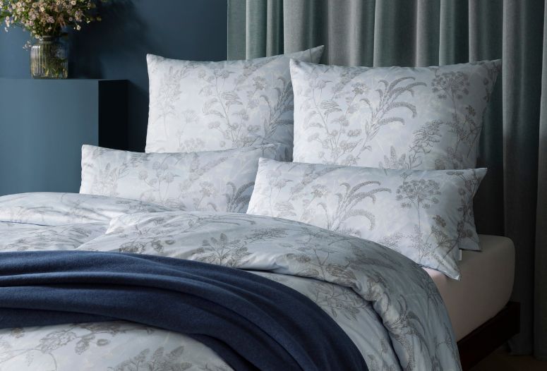 Florale Bettwäsche Elke in hellem Grau-Weiß mit feinem Pflanzenmuster auf modernem Bett, kombiniert mit dunkelblauer Wohndecke Trevi 