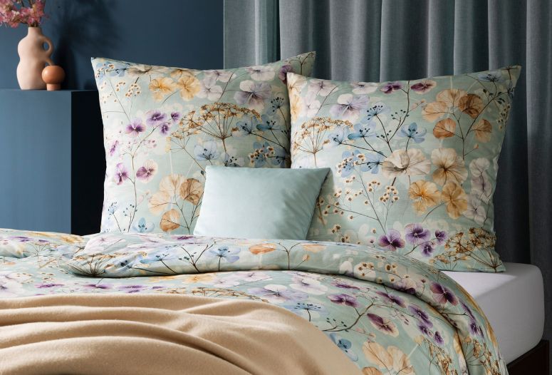 Florale Bettwäsche Sonja in sanftem Grün mit zarten Blütenmotiven auf modernem Bett, kombiniert mit azurfarbenem Uni-Kissen und beigefarbener Wohndecke Trevi