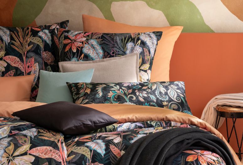 Bettwäsche Juliane mit floralem Blattmuster in bunten Naturtönen kombiniert mit Uni Bettwäsche Valetta in Orange/Apricot, dekoriert mit verschiedenen Dekokissenbezügen und dunkler Wohndecke auf Doppelbett in stilvollem Schlafzimmer