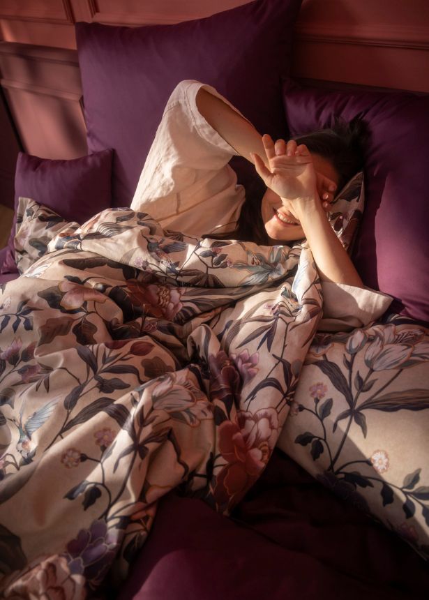 Vom warmen Licht umhüllte Frau in Estella Bettwäsche Pauline in der Farbe Aubergine in gemütlicher Atmosphäre.