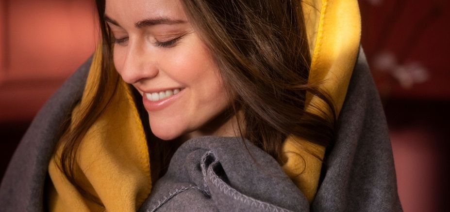 Frau kuschelt sich in Estella Wohndecke Trevi in der Farbe grau und altgold